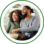 A husband and wife share a bowl of popcorn and sit on the couch.
